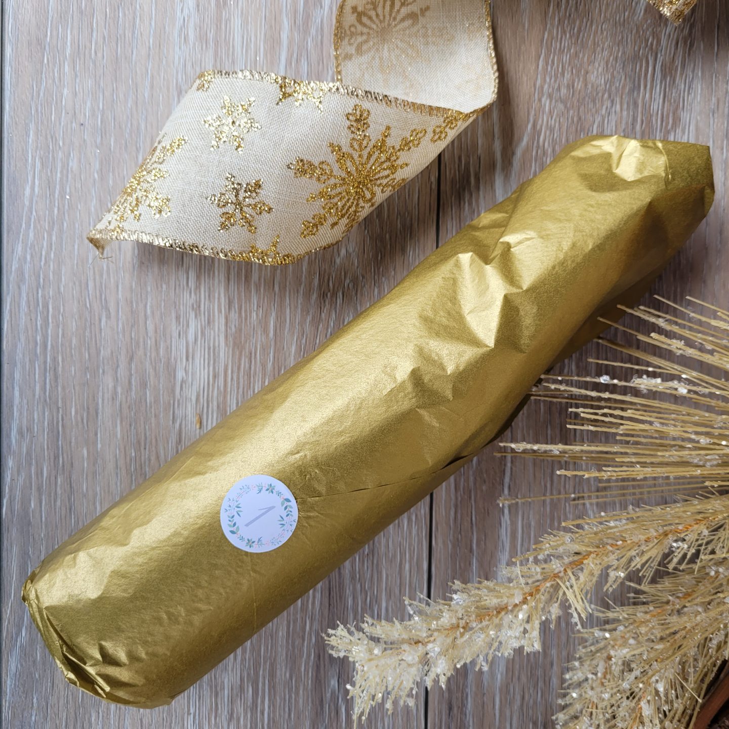 MINI BOTTLE WRAPPED IN GOLD TISSUE, CURL OF A GOLD RIBBON SITS IN FRAME AS WELL AS A GOLD SPRUCE BRANCH