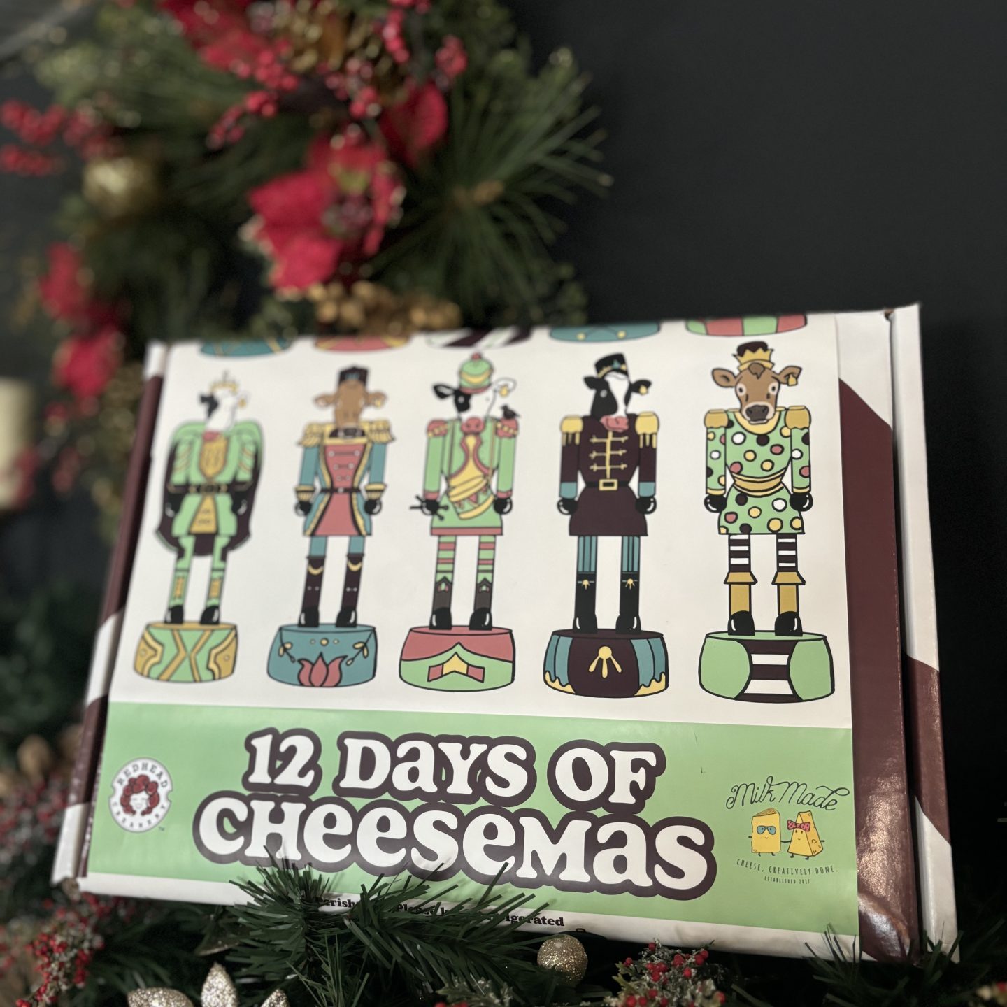 gift box laying in holiday greenery décor/garland, box reads: 12 dats of cheesemas and features illustrated nutcracker cows