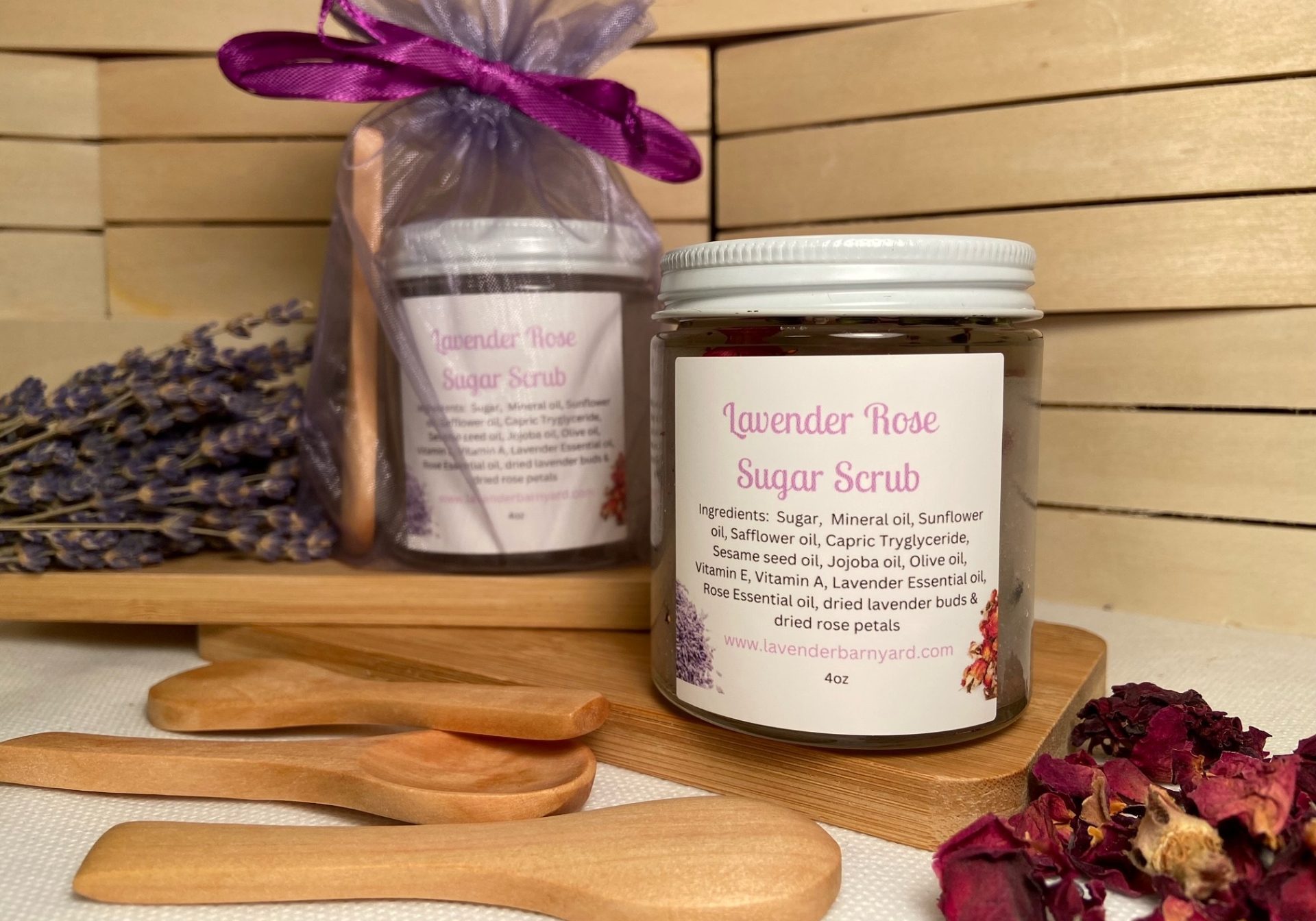 Two small jars or scented scrub pictured with dried lavender and rose petals and small wooden spoons.