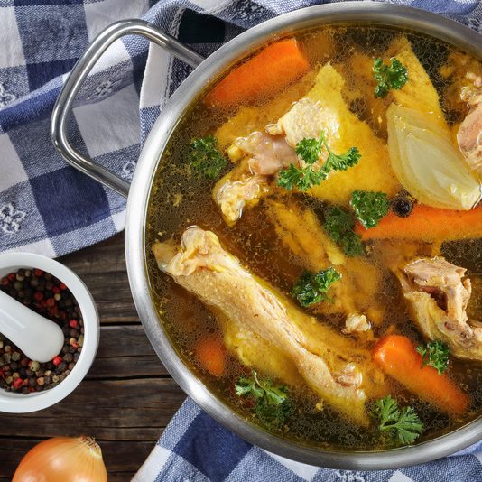 clear Chicken broth with pieces of free range farm organic rooster meat on bone in a metal casserole with kitchen towel and ingredients on wooden table, view from above, close-up